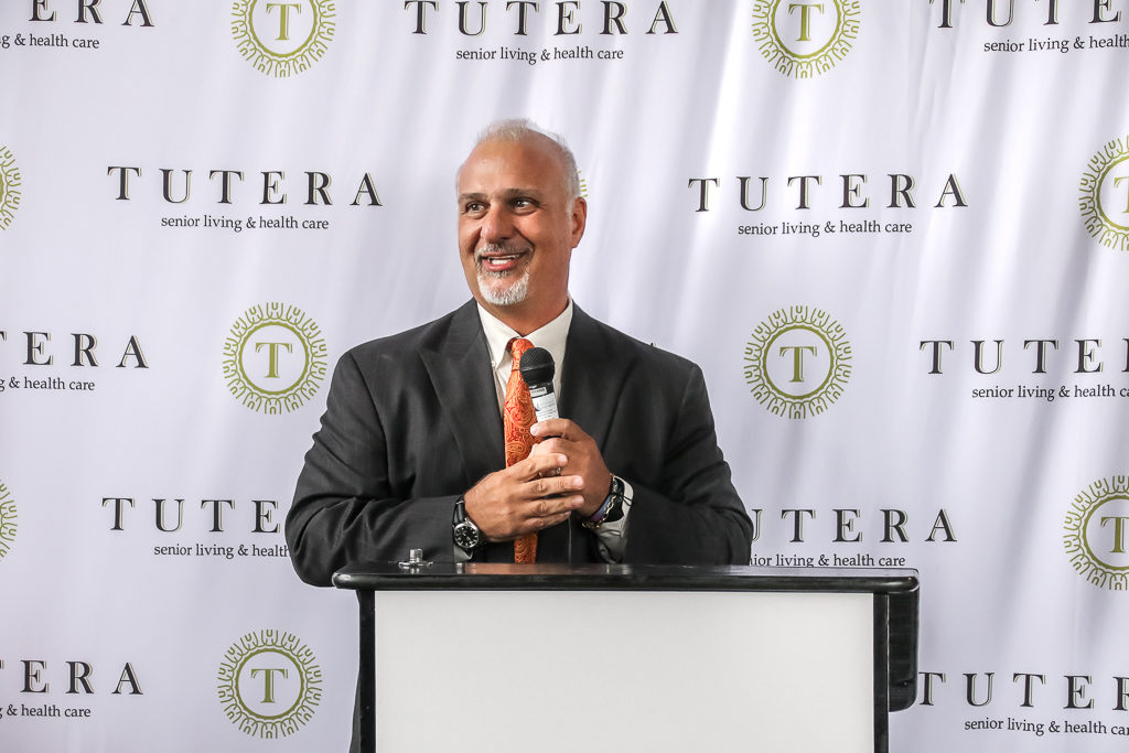 Joe Tutera giving a speech with a microphone in hand, standing at a podium